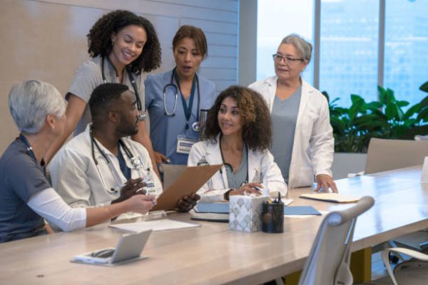 A large group of Doctors, nurses, and other professional medical staff sit closely together as they review a patients case and try to collectively come up with a plan of care.  They are each dressed professionally and focused on the task.