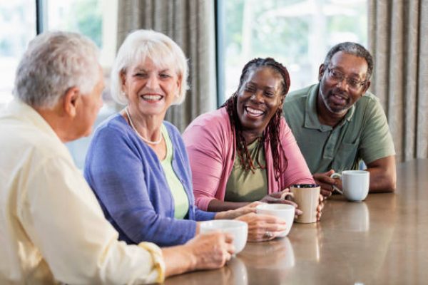 Four multi-ethnic mature and senior adults sitting indoors at a table, drinking coffee, conversing and smiling. The main focus is on an African-American couple in their 50s. Their friends are a senior couple in their 70s.